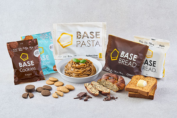 A variety of Basefood products displayed on a tabletop, including packaged cookies, bread, and pasta with some items plated and unwrapped for presentation.