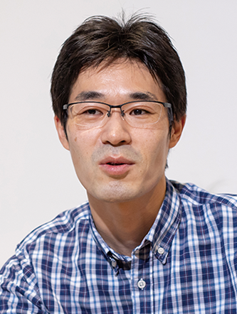 A man wearing glasses and a checkered plaid shirt, looking forward, with a neutral light background.