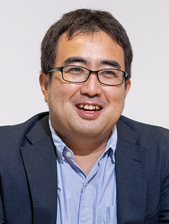 A man wearing glasses and a suit jacket, smiling while looking forward, set against a plain background.