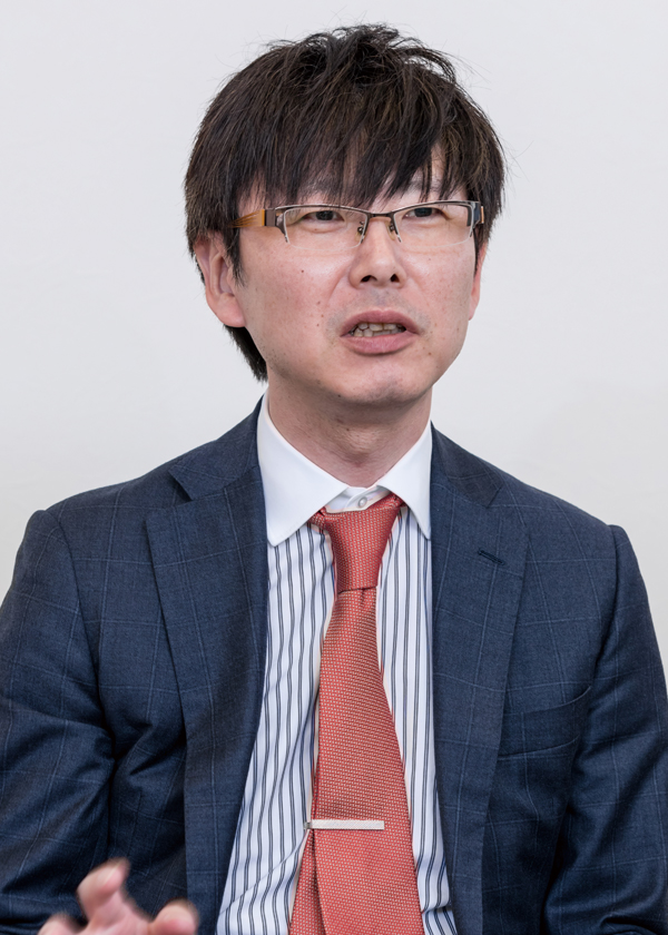A business professional wearing a suit, striped shirt, orange tie, and glasses, pictured in a formal interview portrait against a plain background.