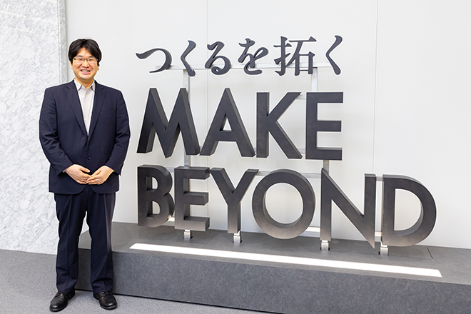 A person in business attire standing beside a large sign displaying the phrase 'MAKE BEYOND' with Japanese text above it, inside an office setting.