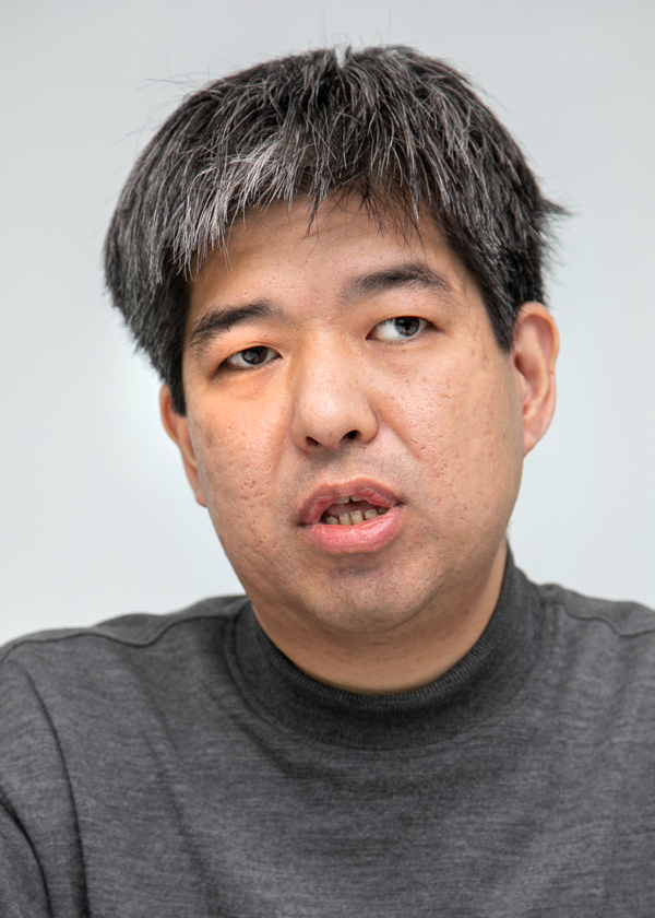 Portrait of a person during an interview, photographed for TV Tokyo. The individual is wearing a dark sweater and is pictured against a neutral background.