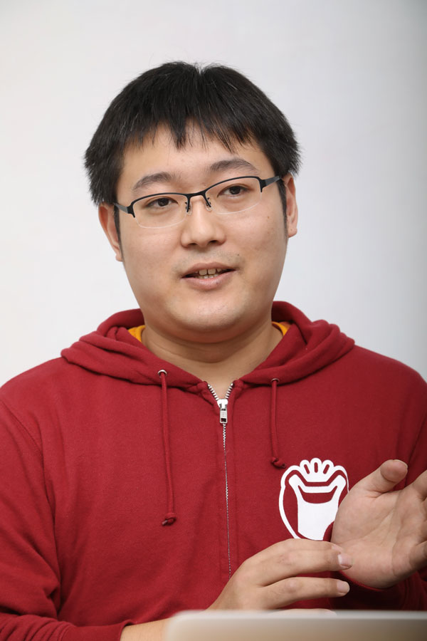 A person wearing glasses and a red hoodie with a white logo is speaking, gesturing with their hands in front of a neutral background.