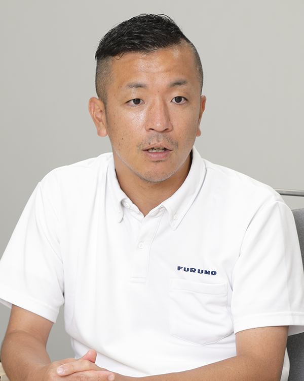 A portrait of a Furuno case study speaker wearing a white polo shirt with the Furuno logo, seated and speaking. Neutral gray background.