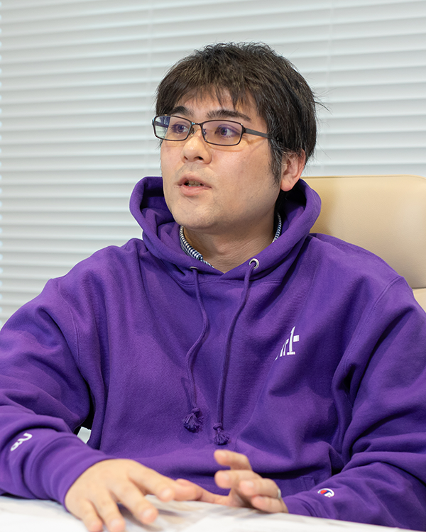 A person wearing a purple hoodie and glasses, speaking or presenting, seated indoors in front of a white blind. Image used for the KDDI Metaverse solution case study on AWS Japan website.
