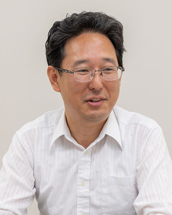 Portrait of a speaker in a white shirt from the Hitachi GLS case study, seated and looking slightly to the side against a neutral background.