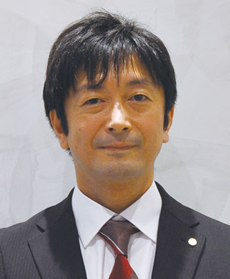A person wearing a dark suit, white shirt, and red tie, with a lapel pin on the jacket.