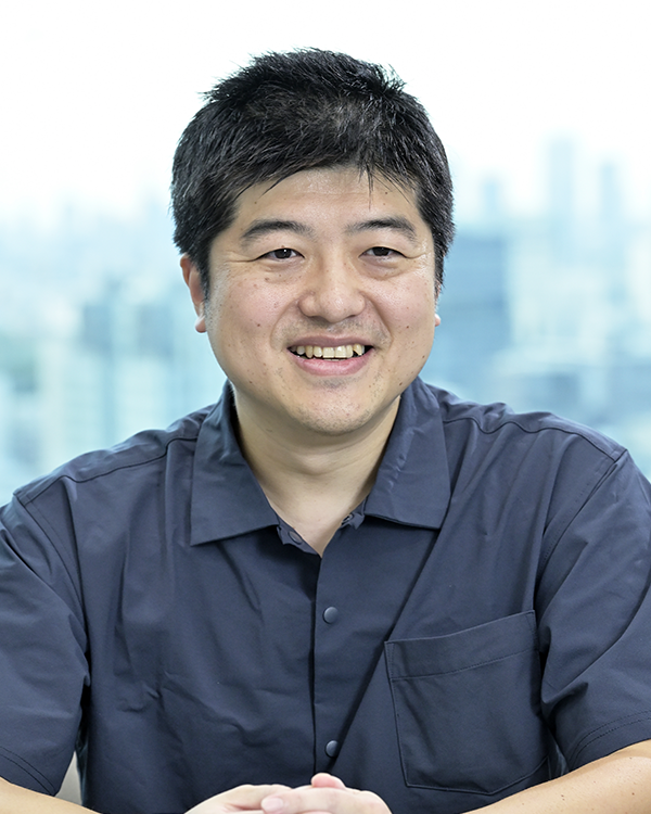A man wearing a dark shirt is smiling while seated in an office environment, with a cityscape visible in the blurred background.