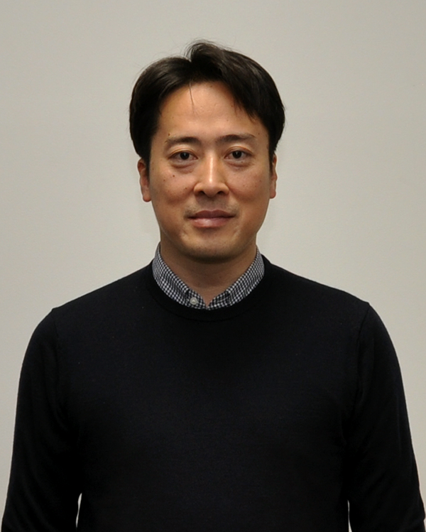 A person wearing a dark sweater over a checkered collared shirt, standing against a plain background.