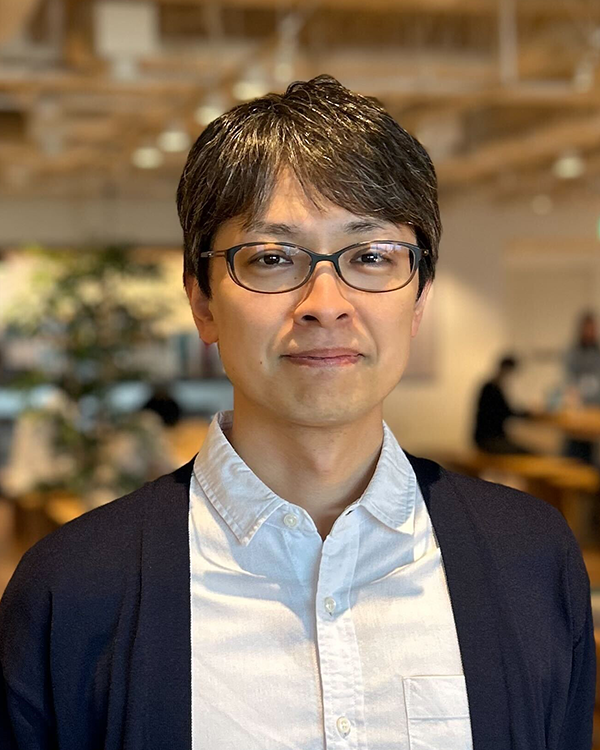 A portrait of an individual wearing glasses and a white shirt with a navy cardigan, standing in a modern office environment. The image is part of a case study for SoftBank, with a blurred background showing people working at desks.
