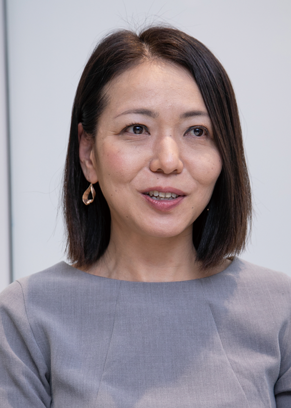 A person with shoulder-length dark hair wearing a gray top and gold earrings.