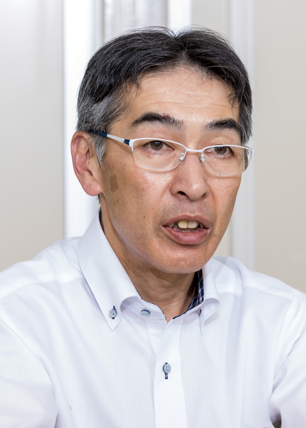 A middle-aged man wearing glasses and a white collared shirt is speaking, photographed indoors in a vertical portrait for an interview.
