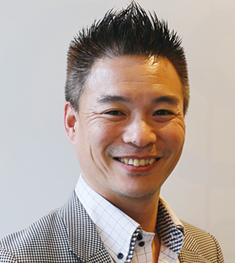 Portrait of a smiling executive from Transcosmos, wearing a checked blazer and white shirt, photographed against a light background.