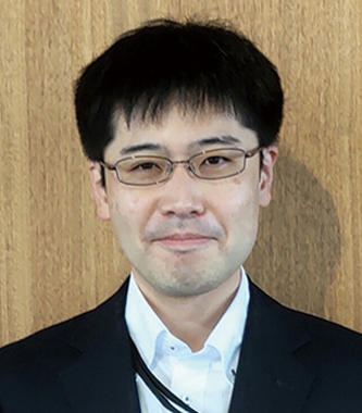 Portrait of a Konica Minolta employee in a suit and glasses standing in front of a wooden background.