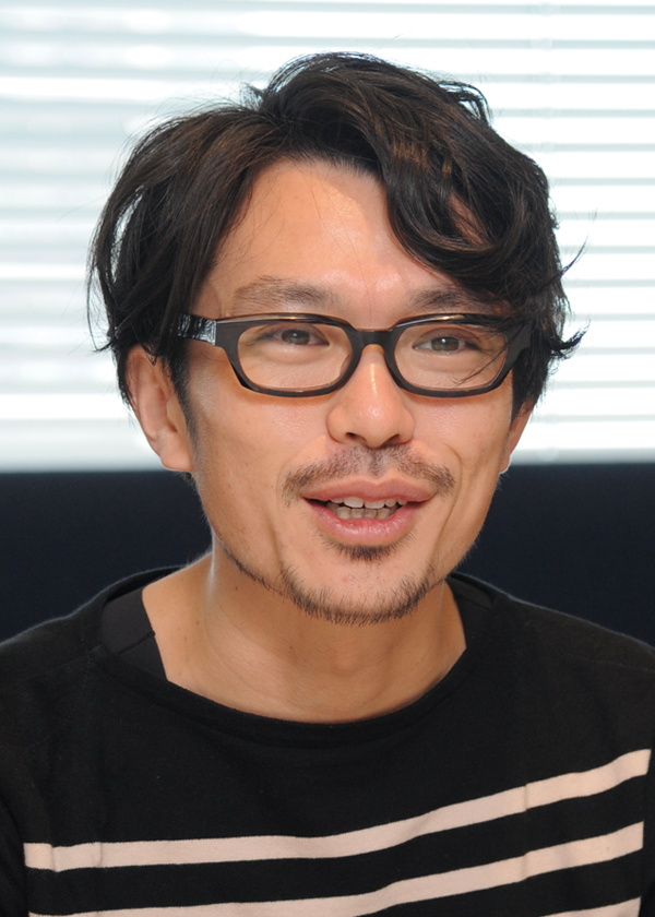 Portrait of an individual wearing glasses and a striped shirt, with short dark hair, sitting in front of a window with blinds.