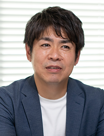 A portrait photograph of Mr. Seike from Desamis, wearing a blue blazer and white shirt, seated in front of a window.