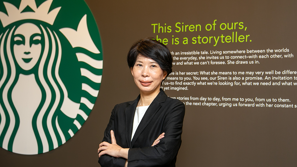A Starbucks Japan executive in business attire stands with arms crossed in front of the Starbucks logo and a wall with the text 'This Siren of ours, she is a storyteller.'