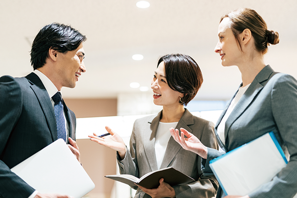 Three business professionals in suits engaged in a discussion, holding documents and laptops in a bright office setting. Represents collaboration and consulting in a professional environment.