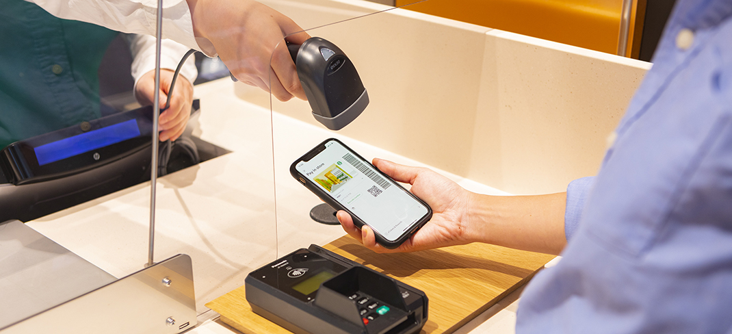 A customer uses a smartphone to make a mobile payment at a Starbucks checkout counter, with a staff member scanning the phone's screen using a barcode reader.