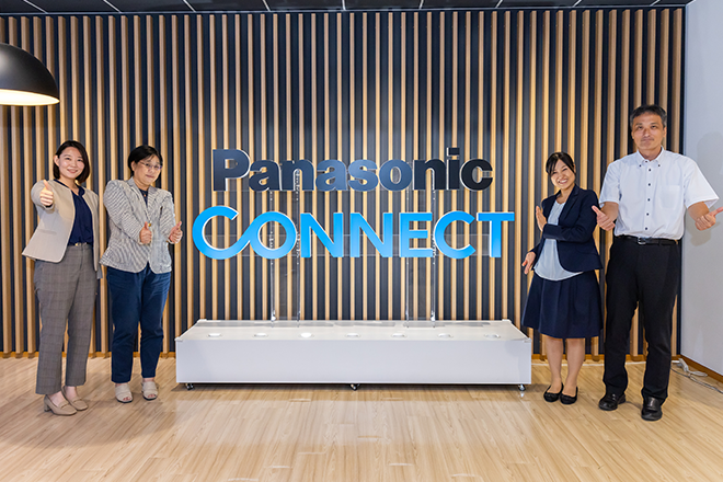 Four people standing and posing in front of a large 'Panasonic Connect' sign at an office with a modern wood panel wall.
