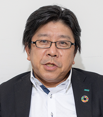 A professional portrait of Mr. Kinoshita wearing glasses and a suit with a pin on his jacket.