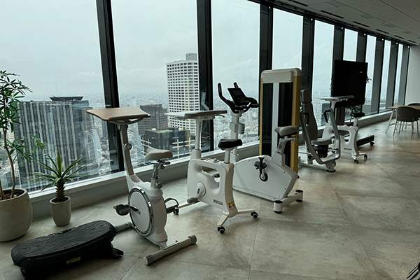 A variety of office fitness equipment, including exercise bikes and desks, arranged by floor-to-ceiling windows with a city view in the background. The space is modern and designed to promote workplace well-being.