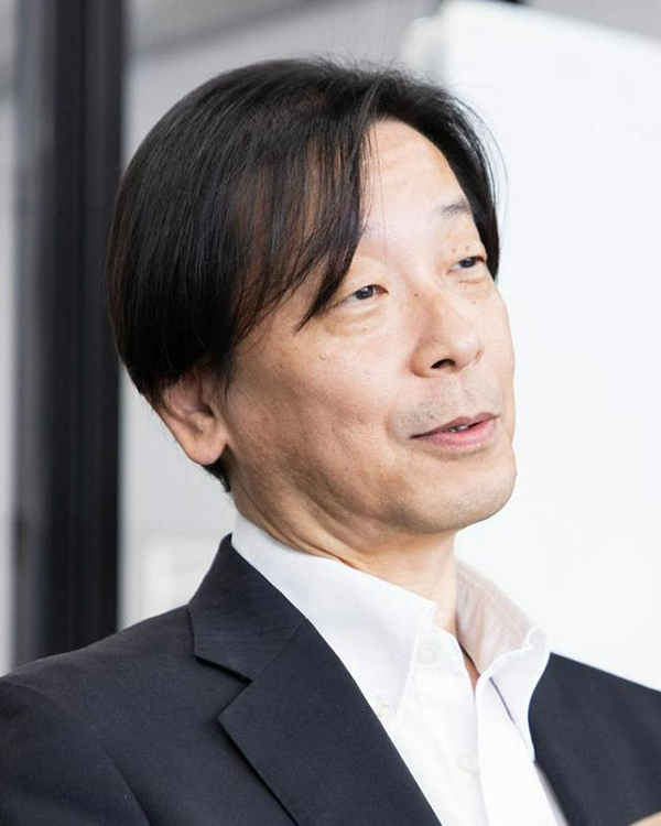 A person wearing a white collared shirt and dark blazer, with short black hair, in an indoor setting.