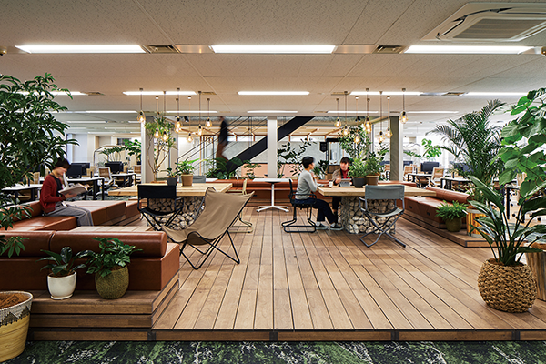 Modern office space with wooden flooring, abundant greenery, lounge seating, and people working at tables.