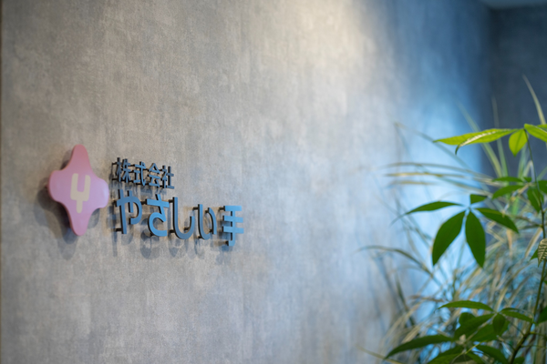 Interior office wall with the Yasashii Te company logo and Japanese text, next to a green plant. The logo is displayed in a calm, professional setting.