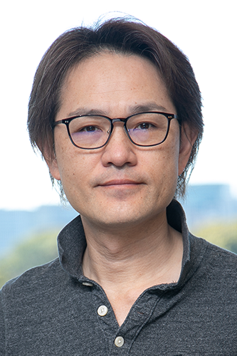 Portrait of a person wearing glasses, grey collared shirt, and a neutral expression, set against a blurred outdoor background.
