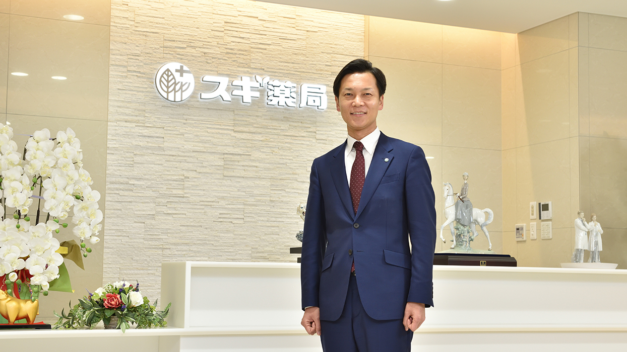 A man in a blue suit standing in a modern office lobby with white orchids, decorative statues, and a sign in Japanese on the wall.