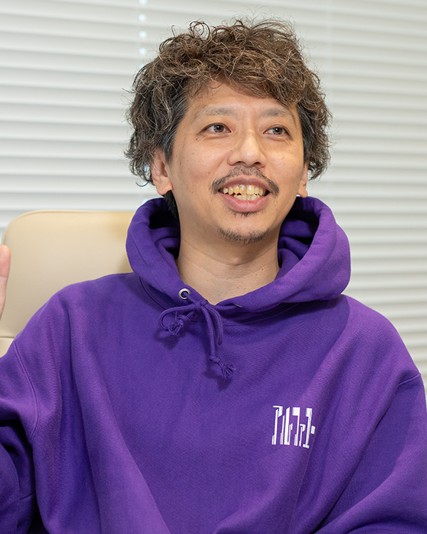 Person wearing a purple hoodie with white text, seated in front of white blinds.