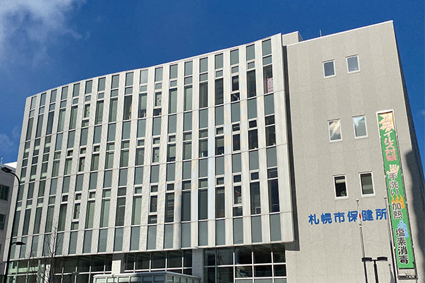 Alt-text: Modern gray building with vertical windows, labeled "Sapporo City Public Health Office" in Japanese, under a clear blue sky.