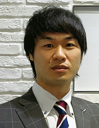 A person wearing a gray plaid suit, white shirt, and striped tie, standing against a white brick wall.