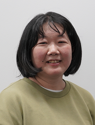 Portrait of a woman with short black hair wearing a green sweater, smiling at the camera against a light background.