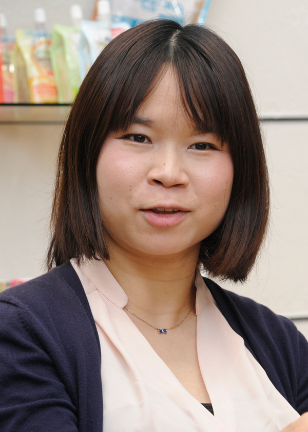 A person wearing a pink blouse, navy cardigan, and a small necklace with a blue pendant, with shelves of colorful products in the background.