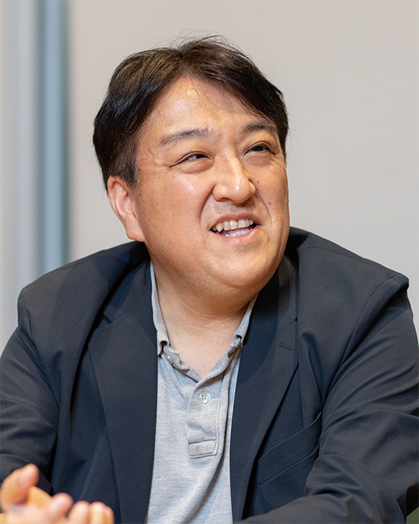 A person wearing a blazer and polo shirt is seated and smiling during an interview, photographed indoors against a neutral background.