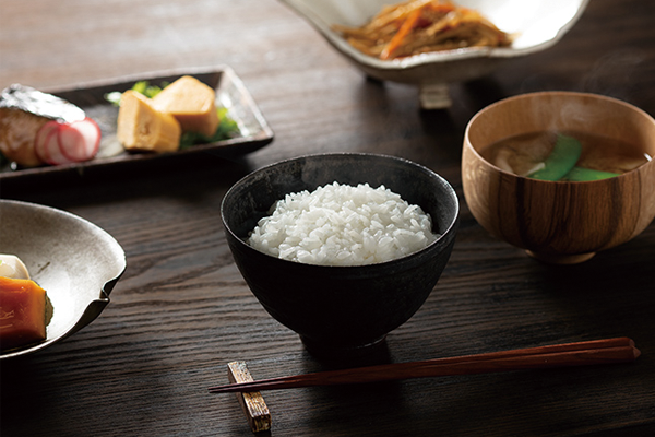 A bowl of steamed white rice served with various traditional Japanese side dishes, including miso soup, pickled vegetables, and egg, presented on a wooden table. Image for the Zojirushi case study on Japanese cuisine.