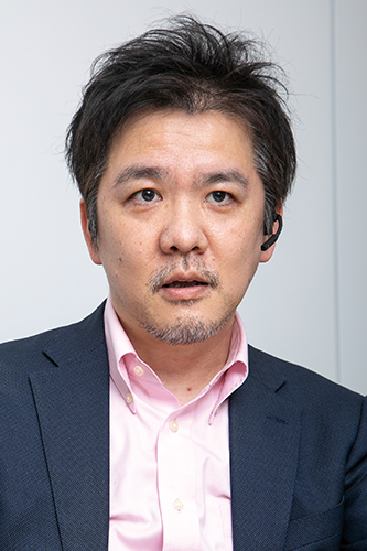 Portrait of a businessman wearing a dark suit, light pink shirt, and headset, looking forward with a neutral expression.
