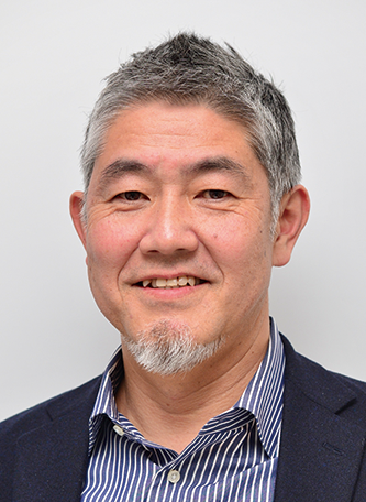 A professional portrait of a smiling middle-aged man with short, graying hair and a goatee, wearing a navy blazer and a striped shirt, against a plain light background.