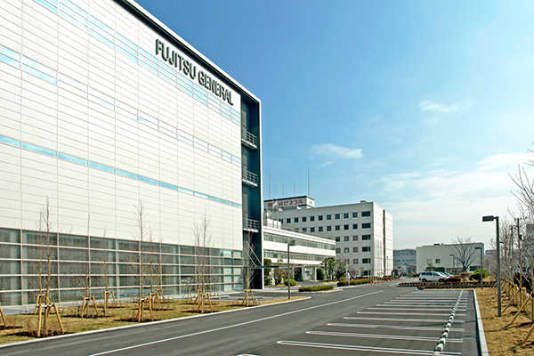 Exterior view of the Fujitsu General headquarters building, showing a modern office facility with a parking lot and clear blue sky.