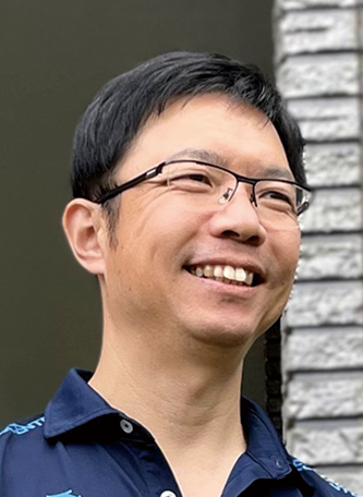 A portrait of a smiling man wearing glasses and a navy shirt, standing in front of a wall with a brick pattern.