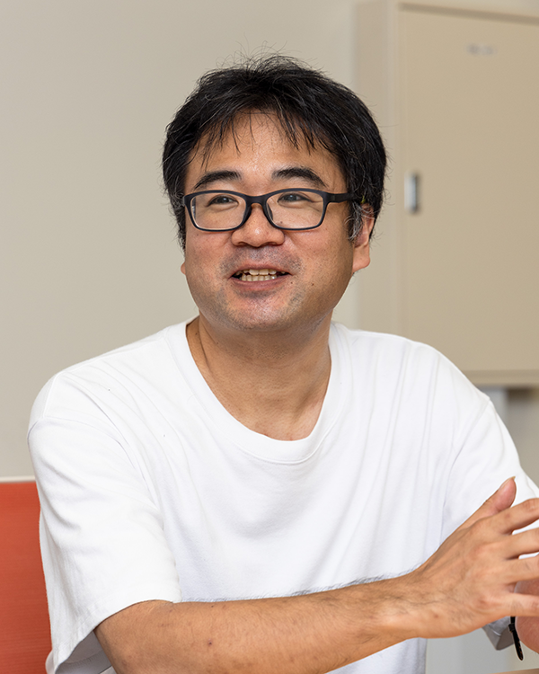 A person wearing a white t-shirt sitting indoors with a beige cabinet in the background.