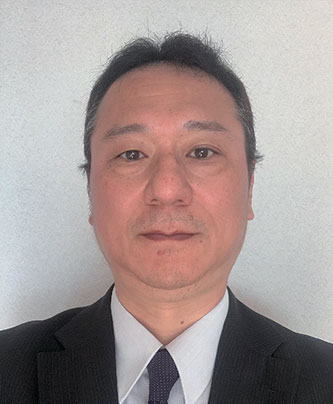 A person wearing a black suit, white shirt, and dark tie against a plain background.
