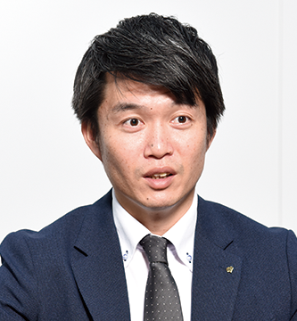 A man in a dark suit and tie, seated and speaking, against a plain light background. Ideal for business or interview context imagery.