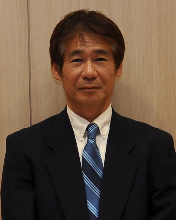 A portrait of a middle-aged man wearing a dark suit, white shirt, and blue striped tie, standing in front of a wooden wall.