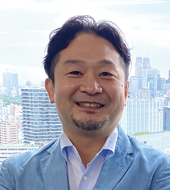 Portrait of an ITOCHU executive smiling, wearing a blue jacket and light blue shirt, standing in front of a large cityscape window background.