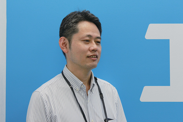 A man in a striped shirt and lanyard speaks during an interview standing in front of a blue background with a partial logo. This image is related to an Airtrip solution case study interview.