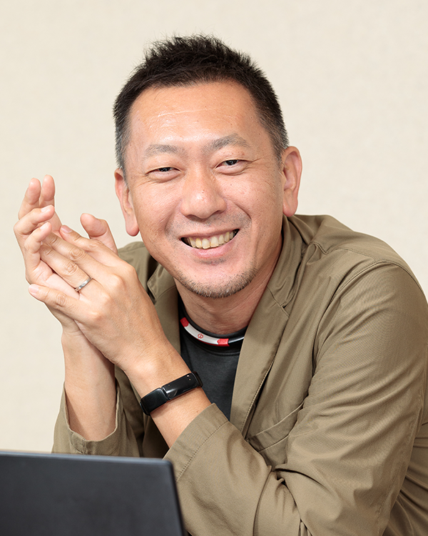 A smiling man in a khaki jacket sits at a desk with his hands clasped, looking at the camera.