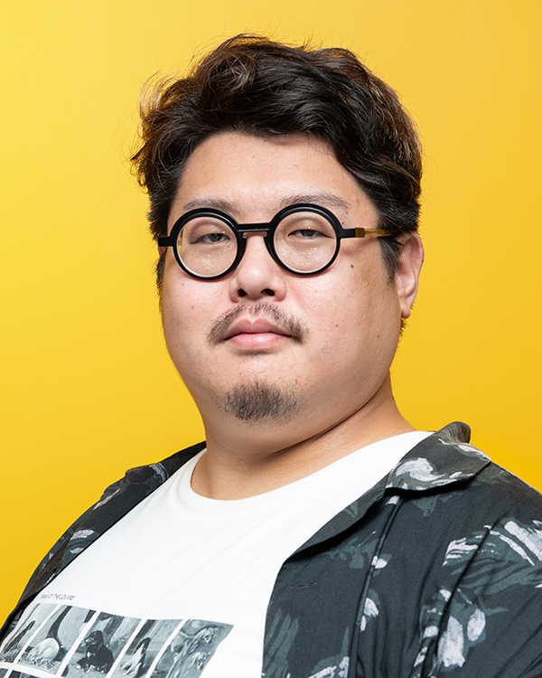 A person wearing a patterned jacket and graphic t-shirt against a bright yellow background.